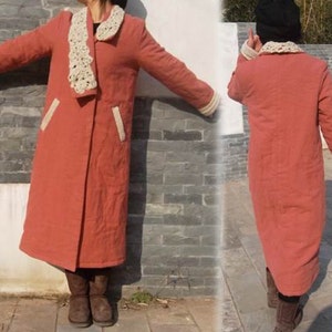 May include: A long, coral-colored coat with a cream-colored crocheted collar and cuffs. The coat features two front pockets with cream-colored trim. The coat is knee-length and has a relaxed fit, suitable for cooler weather.