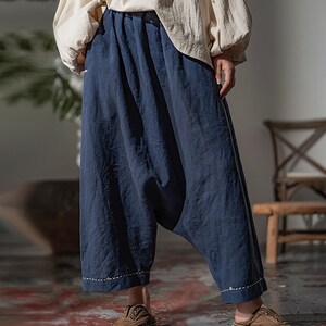 May include: Loose-fitting, navy blue pants with a gathered waist and a relaxed, wide-leg design. The pants have a drop-crotch style and are paired with a cream-colored top. The hemline features a white stitched detail. The person is wearing brown slip-on shoes.