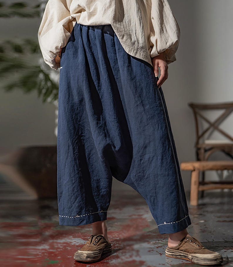 May include: Loose-fitting, navy blue pants with a gathered waist and a relaxed, wide-leg design. The pants have a drop-crotch style and are paired with a cream-colored top. The hemline features a white stitched detail. The person is wearing brown slip-on shoes.