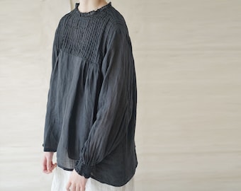 Puede incluir: Una blusa negra de manga larga con cuello fruncido y manga ligeramente abullonada. La blusa está hecha de una tela ligera y transparente.