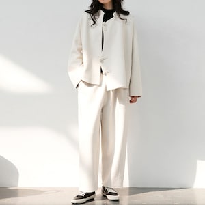 May include: A white linen jacket with a mandarin collar and button closures, paired with matching white linen pants. The model is wearing black shoes.