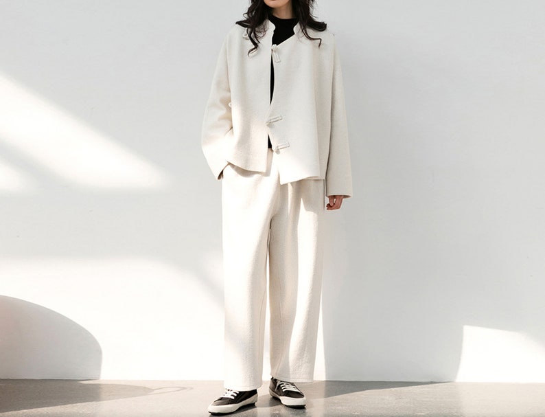 May include: A white linen jacket with a mandarin collar and button closures, paired with matching white linen pants. The model is wearing black shoes.