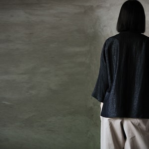 Puede incluir: Una persona vistiendo una blusa de lino negra con superficie texturizada y pantalones beige. La persona est&aacute; de pie frente a una pared gris texturizada.