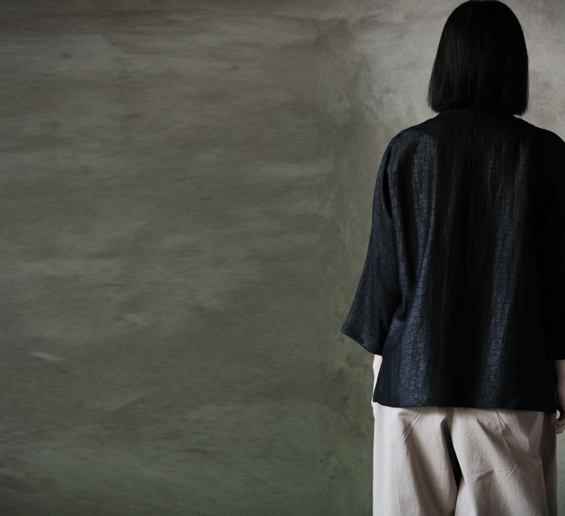 Puede incluir: Una persona vistiendo una blusa de lino negra con superficie texturizada y pantalones beige. La persona est&aacute; de pie frente a una pared gris texturizada.