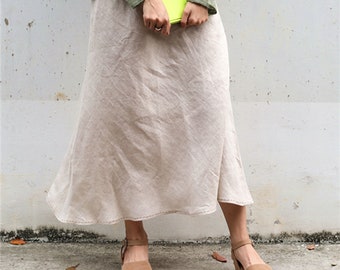 May include: A beige linen skirt with a flared hemline. The skirt is paired with brown sandals. The person is wearing a green long-sleeved shirt and holding a bright green book. The skirt appears to be made of a lightweight fabric.