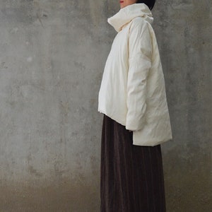 Könnte beinhalten: Cremefarbene, übergroße Jacke mit hohem, rundem Kragen. Die Jacke hat eine lockere Passform und lange Ärmel. Das Model trägt einen langen, dunkelbraunen Rock. Der Hintergrund ist eine schlichte, strukturierte graue Wand.