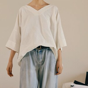 May include: A white, loose-fitting, v-neck blouse with 3/4 sleeves. The blouse is worn with a brown belt and light blue jeans.