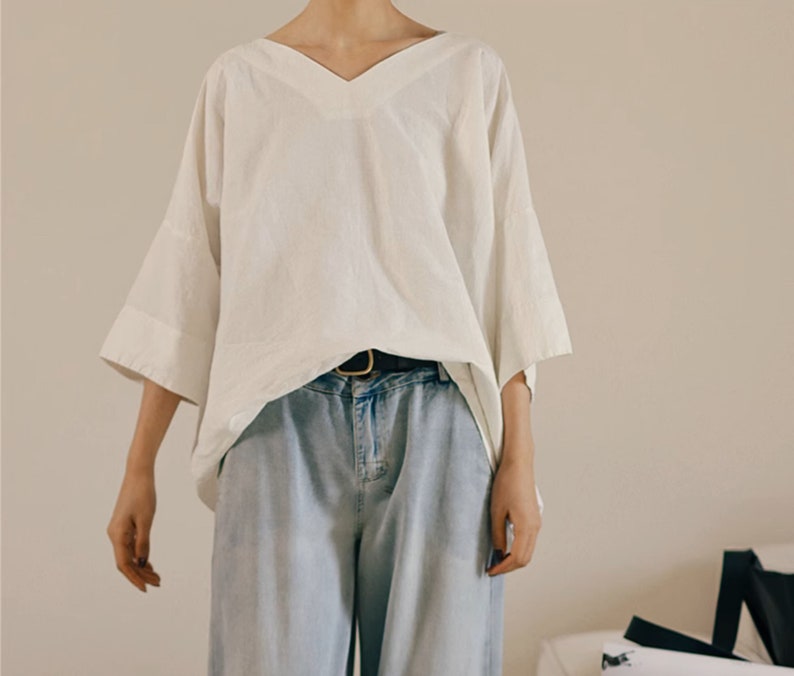 May include: A white, loose-fitting, v-neck blouse with 3/4 sleeves. The blouse is worn with a brown belt and light blue jeans.