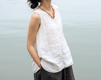 Può includere: Un top senza maniche in lino bianco con scollo a V. Il top ha una vestibilità ampia e un orlo leggermente curvo. La modella indossa una collana marrone e pantaloni grigio scuro. Lo sfondo è un azzurro chiaro sfocato.