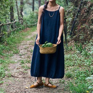 May include: A navy blue, ankle-length dress with spaghetti straps. The dress is worn with brown shoes. A small woven basket filled with green vegetables is held in front of the dress. The background is a forest path.