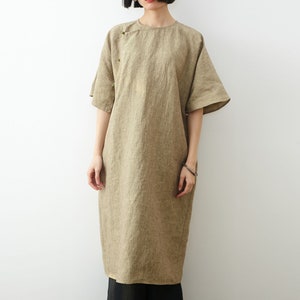 May include: A loose-fitting, knee-length linen dress in a light brown color. The dress has a round neckline and short sleeves. It features a button detail on the left shoulder.