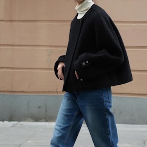 May include: A black, short, wool jacket with a round neckline and buttoned cuffs. The jacket is paired with blue jeans and a white turtleneck. The person is walking on a sidewalk.