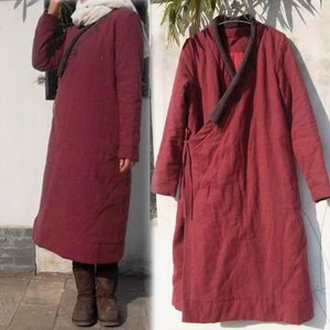 May include: A long, burgundy-colored coat with a wrap-style closure and a dark gray collar. The coat has long sleeves and a tie closure on the side. The coat is made of a quilted material and appears to be warm.