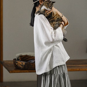 May include: A woman wearing a white linen blouse and a striped skirt holds a tabby cat in her arms. She is wearing a hat and has her hair pulled back.