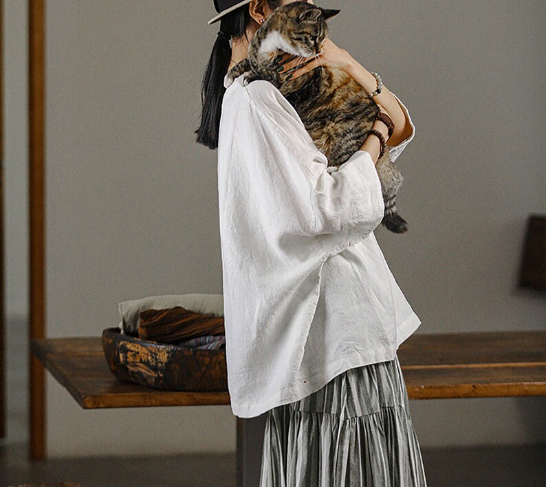 May include: A woman wearing a white linen blouse and a striped skirt holds a tabby cat in her arms. She is wearing a hat and has her hair pulled back.