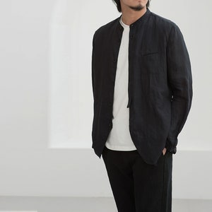 May include: Black linen shirt with a mandarin collar and a single chest pocket. The shirt is worn over a white t-shirt and paired with black pants. The outfit is simple and casual, suitable for everyday wear.