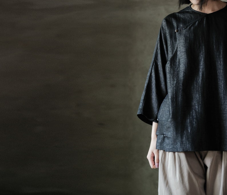 Puede incluir: Una blusa negra holgada con un escote ligeramente asim&eacute;trico y un detalle de bot&oacute;n &uacute;nico. La blusa est&aacute; hecha de una tela texturizada y se lleva con pantalones beige.