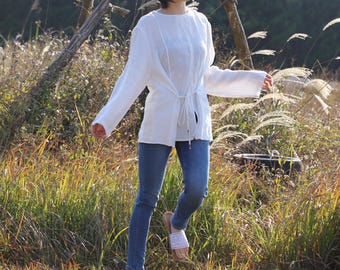 May include: A white long-sleeve top with a drawstring waist and blue jeans. The top has a round neckline and the jeans are a classic denim style. The person is wearing white sandals with a woven design.