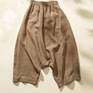May include: Brown linen pants with an elastic waistband and a relaxed fit. The pants have a drop crotch design and are finished with white stitching along the hems. The fabric has a slightly textured appearance.