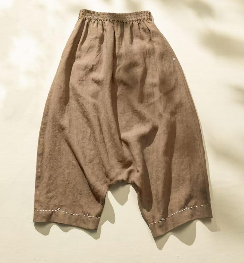 May include: Brown linen pants with an elastic waistband and a relaxed fit. The pants have a drop crotch design and are finished with white stitching along the hems. The fabric has a slightly textured appearance.
