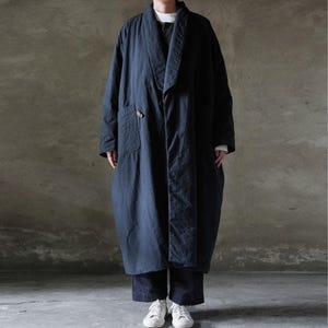 Peut inclure: Long manteau bleu marine avec col châle et fermeture à bouton unique. Une poche matelassée est visible sur le côté gauche. Le manteau est porté avec un pantalon sombre et des baskets blanches. Style minimaliste et décontracté.