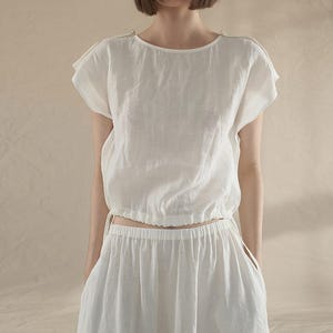 May include: A white linen top with a gathered elastic waistband and short sleeves. The top is paired with a matching white linen skirt.