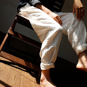 May include: A person wearing white linen trousers with the cuffs rolled up, sitting on a wooden chair. The person's feet are bare and the floor is made of wood.
