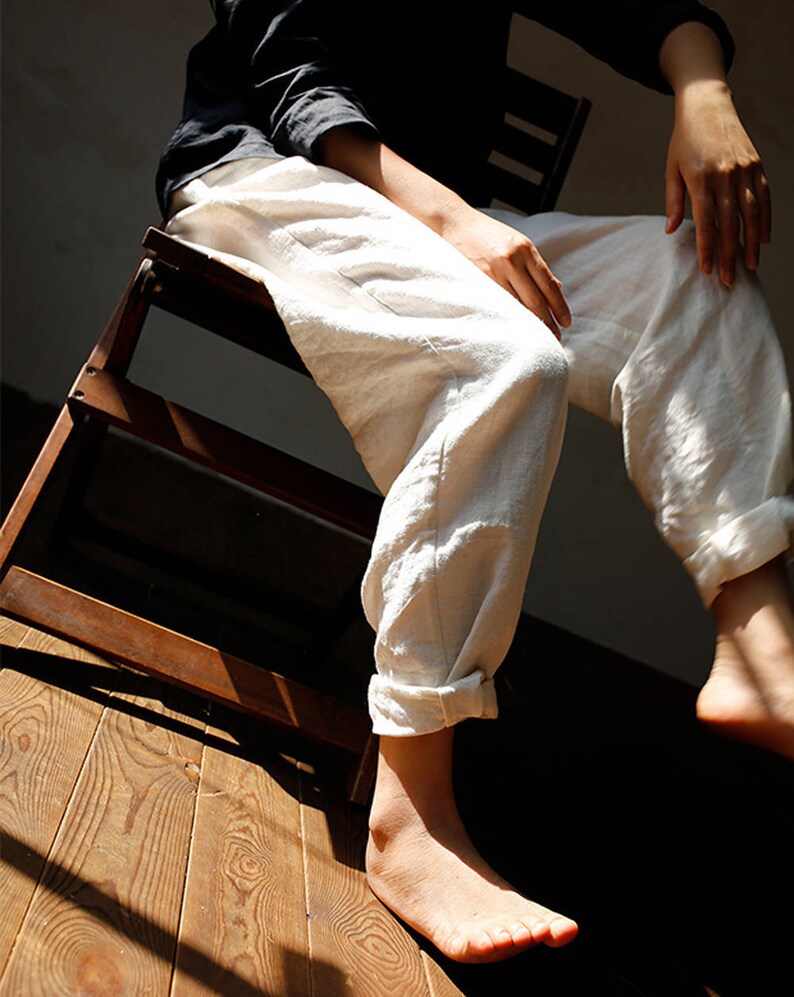 May include: A person wearing white linen trousers with the cuffs rolled up, sitting on a wooden chair. The person's feet are bare and the floor is made of wood.