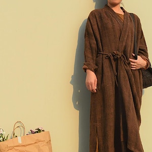 May include: A long brown robe with a tie belt. The robe is made of a soft, textured fabric. The person is wearing the robe and standing in front of a light yellow wall.