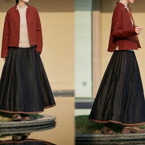 May include: A two-piece outfit featuring a brick-red quilted jacket and a long, dark denim skirt. The skirt has a wide, flowing silhouette with brown trim. The jacket is buttoned and worn over a light-colored top. The outfit is paired with brown shoes.