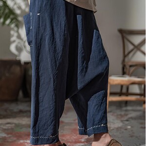 May include: Navy blue linen pants with a relaxed fit and white stitching details. The pants have a wide leg design and a single pocket. The model is wearing brown slip-on shoes. The pants are part of a casual clothing collection.
