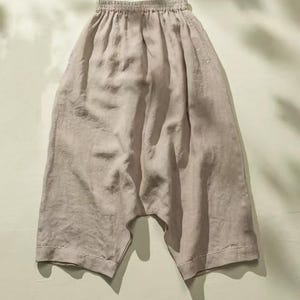 May include: Light beige linen pants with an elastic waistband and wide, cropped legs. The pants have a relaxed fit and are made of a soft, flowing fabric. The hemline features a decorative white stitching detail. The pants are suitable for casual wear.