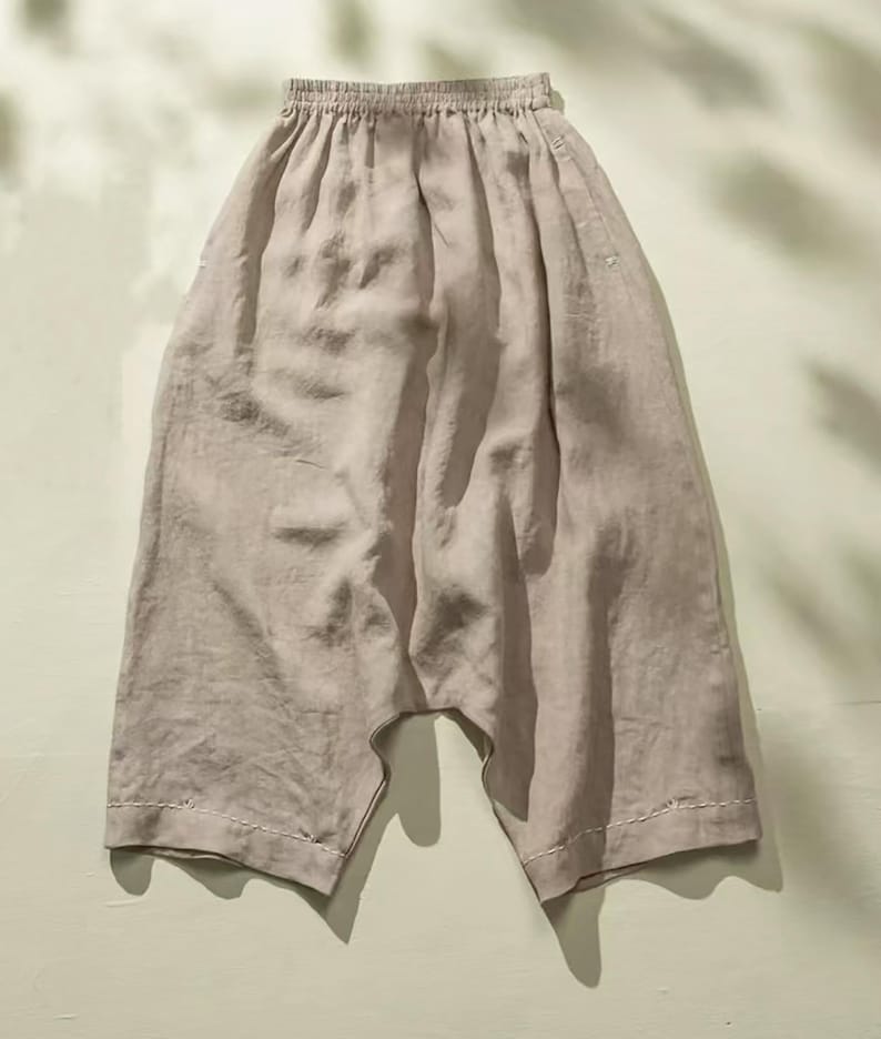 May include: Light beige linen pants with an elastic waistband and wide, cropped legs. The pants have a relaxed fit and are made of a soft, flowing fabric. The hemline features a decorative white stitching detail. The pants are suitable for casual wear.