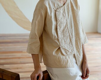 May include: Beige linen top with three-quarter sleeves and a unique asymmetrical design. The top features a wrap-style closure with a button detail and is paired with white pants. The fabric has a textured appearance, suggesting a natural fiber.