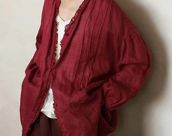 May include: A burgundy-colored, long-sleeved blouse with a ruffled trim along the neckline and front. The blouse has a button-down closure and vertical pleats on the front. The sleeves are gathered at the cuffs, creating a voluminous shape. A white top is visible underneath.