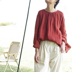 May include: A woman wearing a loose-fitting, rust-colored linen top with a gathered neckline and long sleeves. She is also wearing white, wide-leg pants.