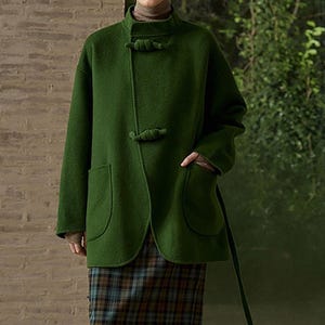 May include: A green wool coat with a mandarin collar and toggle fastenings. The coat has two front pockets and a long, attached belt. The coat is worn over a plaid skirt. The coat's colour is a deep, forest green.