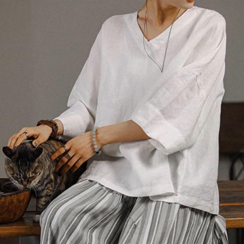 May include: A white linen blouse with a v-neck and 3/4 sleeves. The blouse is loose fitting and has a relaxed, casual style. The model is wearing the blouse with a striped skirt and is holding a cat.