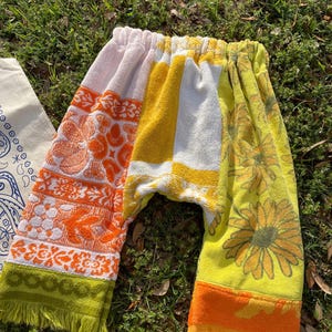 May include: Colorful patchwork pants made from repurposed towels. The pants feature sections of pink, orange, yellow, and white, with floral and geometric patterns. The bottoms have fringe in green and orange. The pants are laying on green grass.