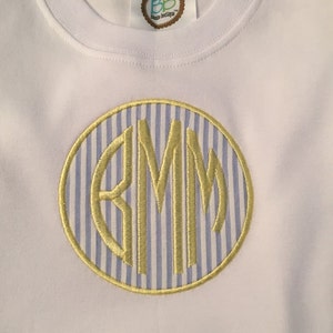 May include: White baby onesie with a yellow embroidered monogram in a circle. The monogram is "BMM" and the circle has a blue and white striped pattern.