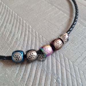 May include: A black leather cord necklace with six silver beads. Each bead has a yin yang symbol engraved on it. The beads are in different colors, including blue, silver, rainbow, brown, and black.