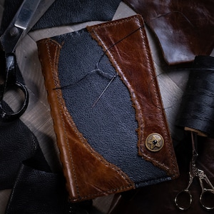 May include: A handcrafted leather journal with a two-tone design. The journal has a dark blue central panel framed by distressed brown leather. A needle and thread are visible, along with a star-shaped button.