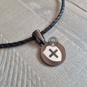 May include: A black leather braided necklace with a black metal pendant featuring a silver shield with a white cross.