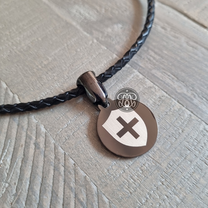 May include: A black leather braided necklace with a black metal pendant featuring a silver shield with a white cross.