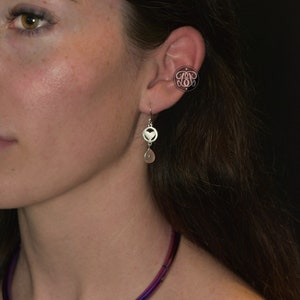 May include: Silver dangle earrings with a heart design and a teardrop-shaped gemstone. A matching ear cuff with an ornate design is also visible. The jewelry is worn on the ear of a person with brown hair.