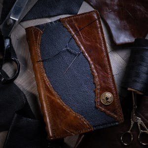 May include: A dark blue leather journal with brown leather accents and a star-shaped metal button. The journal is being stitched together with a needle and thread.