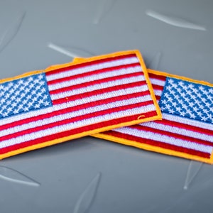 May include: Two small American flag patches with yellow borders. The flags are made of fabric and have a stitched design.