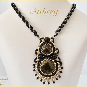 Op de afbeelding: Een zwarte en gouden kralenketting met een grote, ingewikkelde hanger met een zwart en gouden patroon. De hanger is gemaakt van meerdere lagen kralen en heeft een geometrische vorm. De ketting heet "Aubrey".