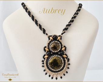May include: A black and gold beaded necklace with a large, intricate pendant featuring a black and gold patterned design. The pendant is made up of multiple layers of beads and has a geometric shape. The necklace is called "Aubrey".