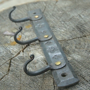 May include: Hand-forged metal wall hook with three curved hooks. The dark gray metal has decorative floral accents and is secured with brass-colored rivets. The hook is mounted on a wooden surface.